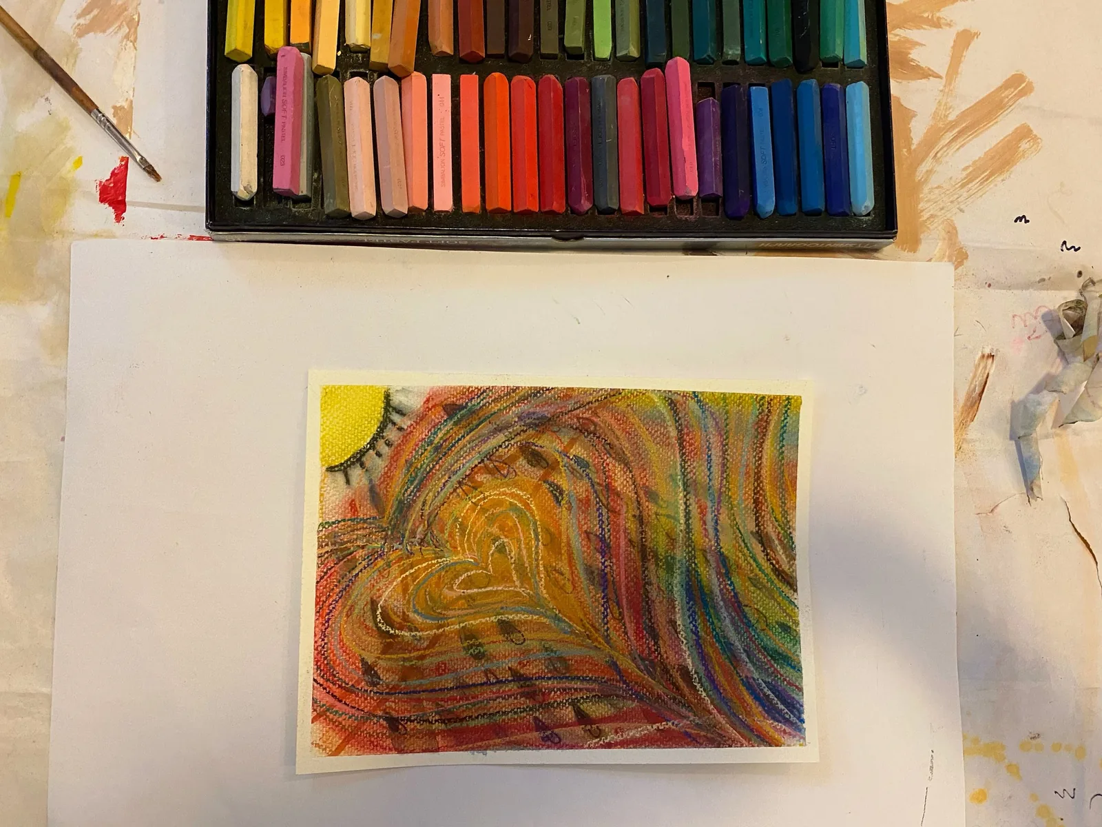 粉彩畫的愛心與陽光，暖色線條層層環繞出一顆心，桌上擺著整排粉彩條